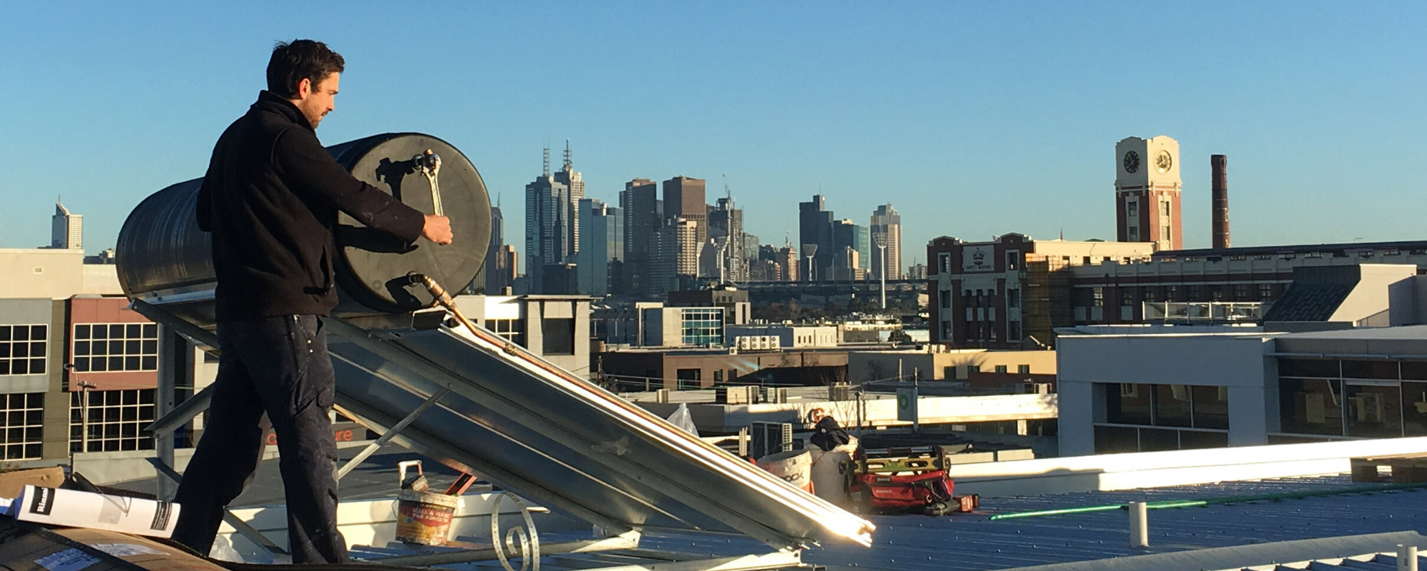 Plumber working on Solar Hot water unit on roof in front of Melbourne skyline